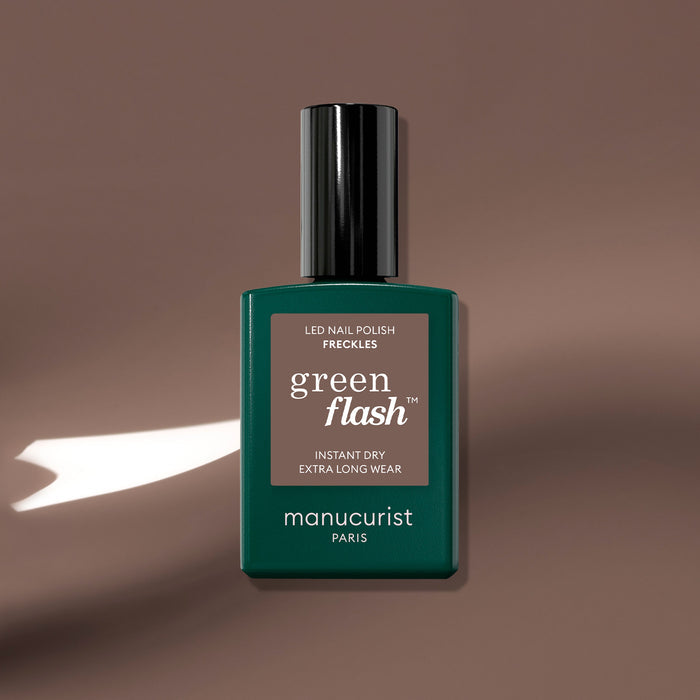 A bottle of Manucurist Freckles gel polish sits on a smooth, wood-toned brown surface with a white light shadow. The label reads: Freckles, Instant Dry, Extra Long Wear.