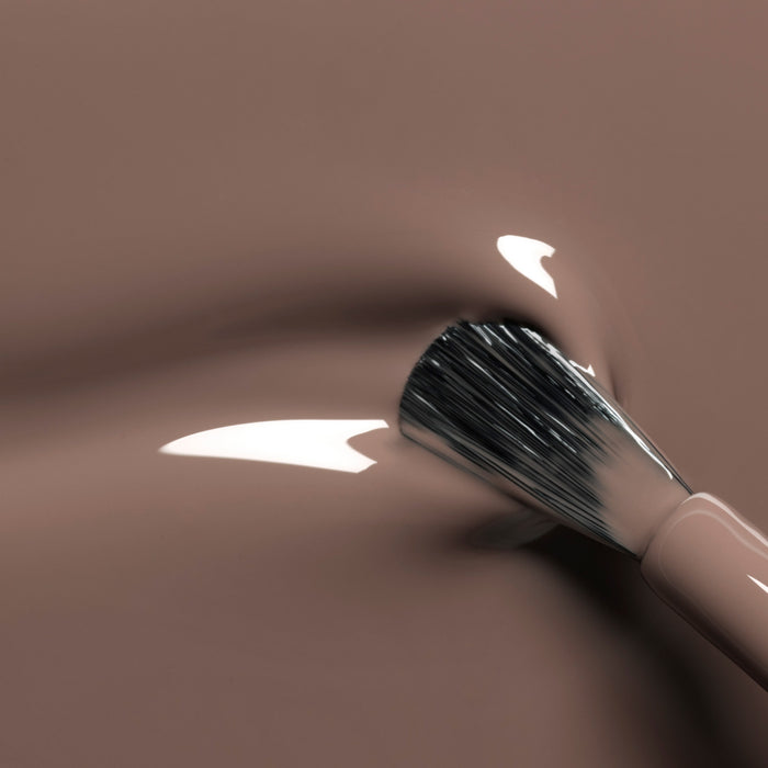 A close-up of a brush applying Manucurist's Freckles, a glossy wood-toned brown gel polish, showing smooth, shiny strokes and reflections on the surface.