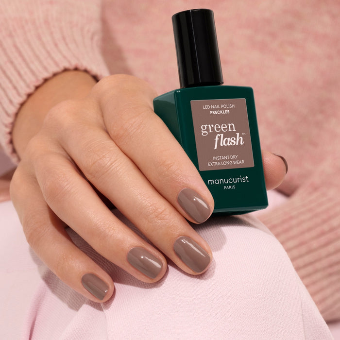 A hand with neatly manicured nails painted in the wood-toned brown shade Freckles by Manucurist holds a green bottle of Green Flash LED gel polish. The person is dressed in light pink clothing.