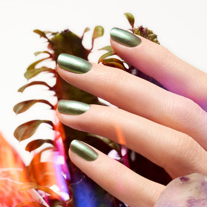 A close-up of a hand with Manucurist Groove metallic green gel polish, fingers gently resting on leafy foliage amid green and reddish leaves softly lit in the background.