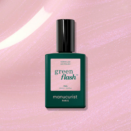 A bottle of Manucurist Halo gel polish stands upright on a shimmery pink background. The dark green bottle with a pale pink label features the product name "Halo" and the Manucurist brand for this innovative LED nail polish.
