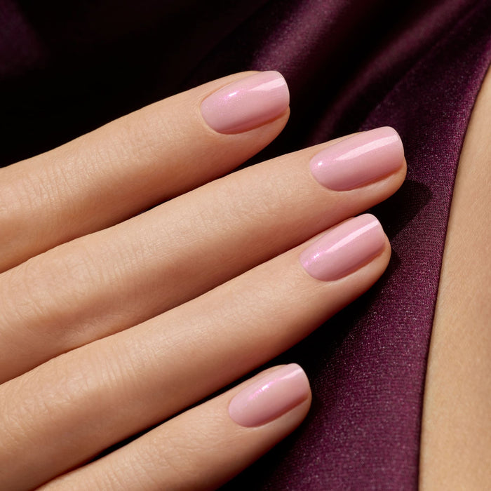 A close-up of a hand with neatly manicured nails painted in Manucurist’s Halo, a glossy light pink milky mauve gel polish, resting on smooth deep purple satin.
