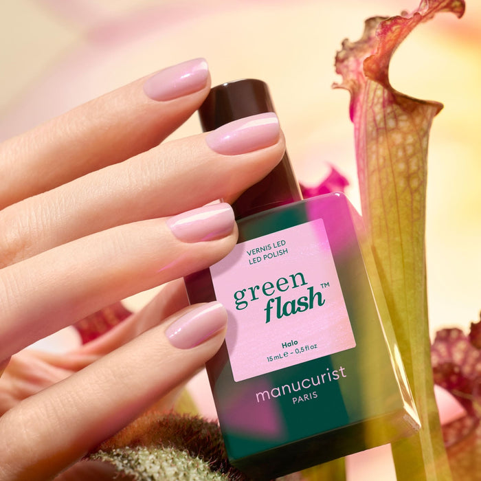 A hand with glossy, pale pink nails holds a bottle of Halo gel polish by Manucurist, with carnivorous plants in the background.