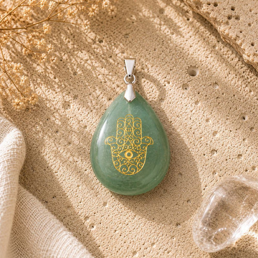A green teardrop pendant with a gold Hamsa hand design and eye motif, resting on a textured beige stone surface with dried flowers and a clear crystal nearby.