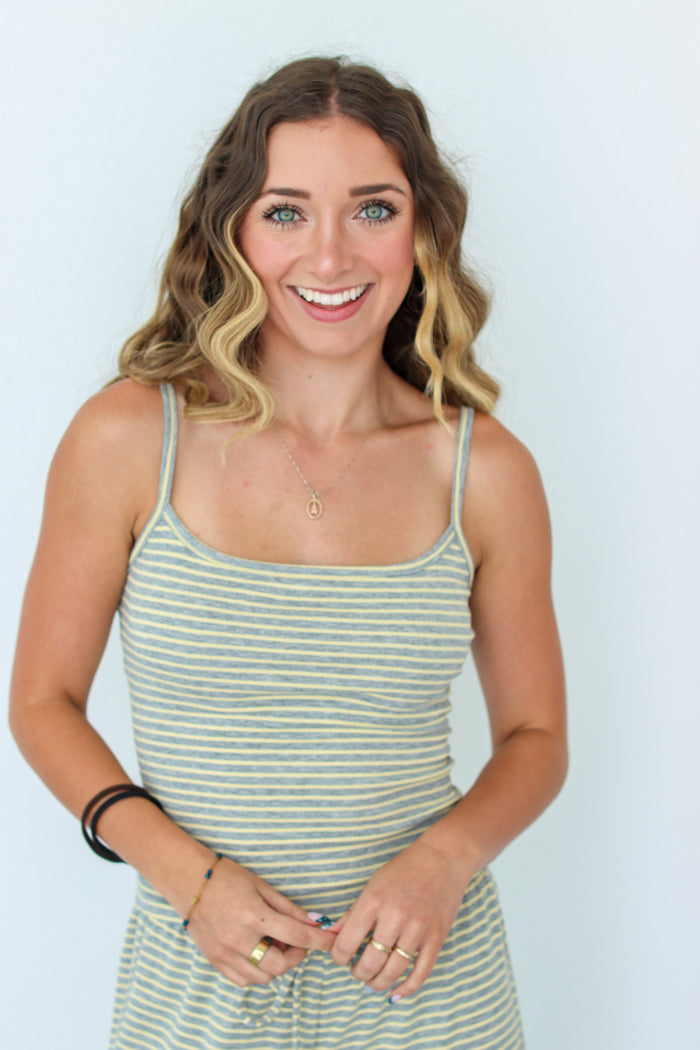 A smiling woman with wavy blonde-highlighted hair wearing a light sage green and yellow striped spaghetti-strap tank dress, standing against a plain light blue background.
