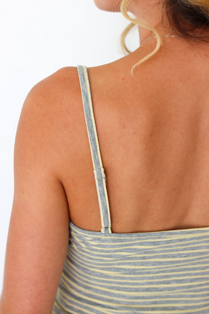 Close-up back view of a woman wearing a striped camisole top with thin gray and yellow straps against a white background.