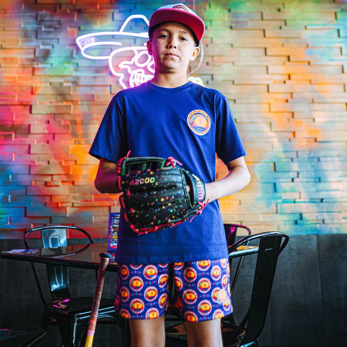 Child holding a baseball glove in front of a colorful brick wall wearing purple taco shorts.