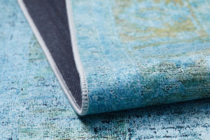 A close-up of the Imperial Jewel - Washable Rug - JR162 in blue and green, ideal for high-traffic areas, shows one corner folded to reveal its dark non-slip backing and white stitched edge.