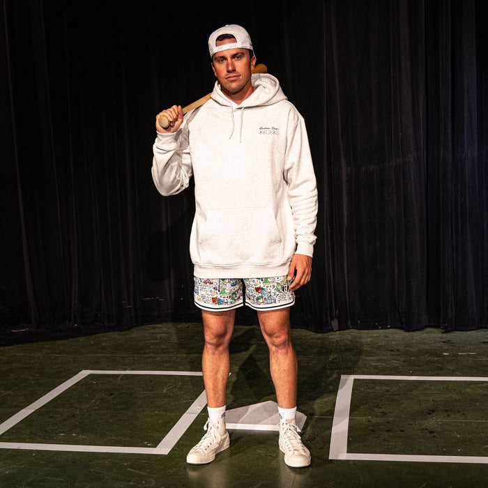 Adult male model holding a bat over his shoulder on a stage set, wearing the white Jackson Olson x BL101 Hometown Shorts with all-over illustrated print paired with a heather gray hoodie