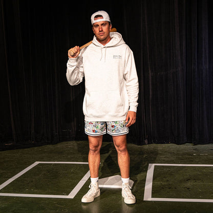Adult male model holding a bat over his shoulder on a stage set, wearing the white Jackson Olson x BL101 Hometown Shorts with all-over illustrated print paired with a heather gray hoodie