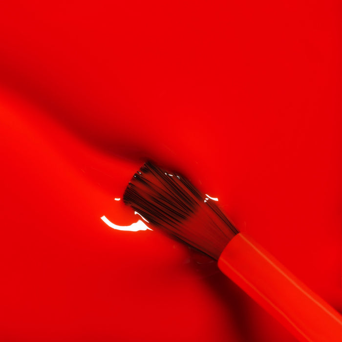 A close-up of a paintbrush with orange bristles dipped in Manucurist's Lava Gel Polish, laying down glossy, pop red strokes and reflections—perfect for capturing the vibrant mood of long summer nights.