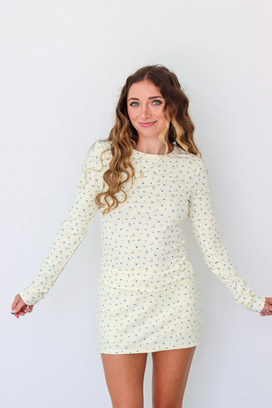 A woman modeling a long-sleeve mini dress with a small, dense all-over print in a light cream or off-white color. The dress has a crew neck and is shown against a plain white studio background.