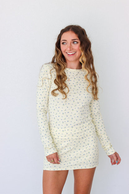 A woman modeling a long-sleeve mini dress with a small, dense all-over print in a light cream or off-white color. The dress has a crew neck and is shown against a plain white studio background.