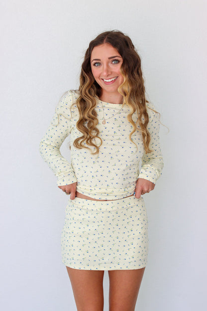 A woman modeling a long-sleeve mini dress with a small, dense all-over print in a light cream or off-white color. The dress has a crew neck and is shown against a plain white studio background.