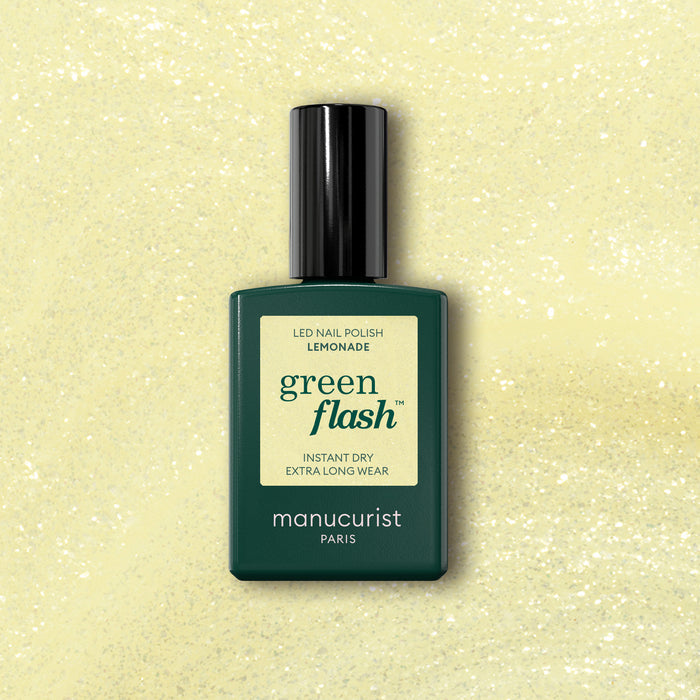 A bottle of Manucurist Lemonade gel polish stands against a yellow glittery background, with a dark green bottle, pale yellow label, and black cap—showcasing the brand's light yellow shade.