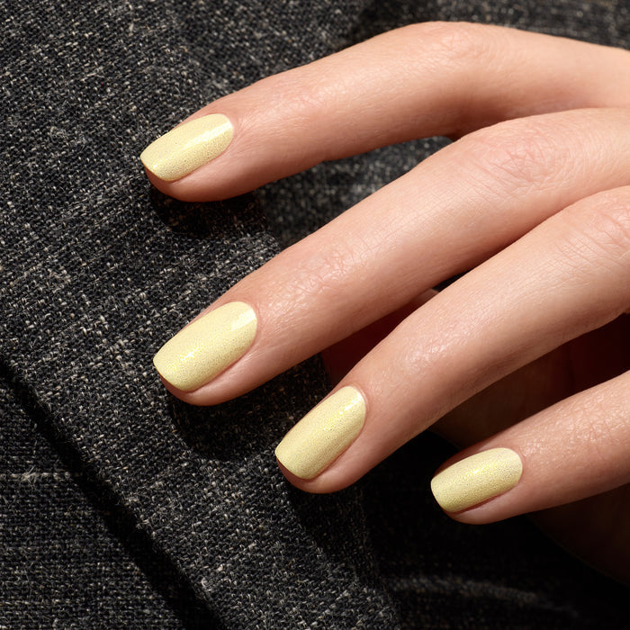 A close-up of neatly manicured nails painted with Manucurist's Lemonade, a shimmery pale yellow gel polish, resting on textured dark gray fabric.
