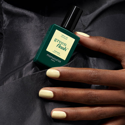A hand with smooth, pale yellow gel polished nails holds a bottle of Manucurist Lemonade nail polish against a dark, shiny fabric background.