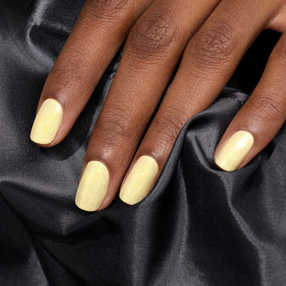 A hand with smooth brown skin displays neatly manicured nails painted in Manucurist's Lemonade, a soft, shimmery pale yellow gel polish, resting on black satin fabric.
