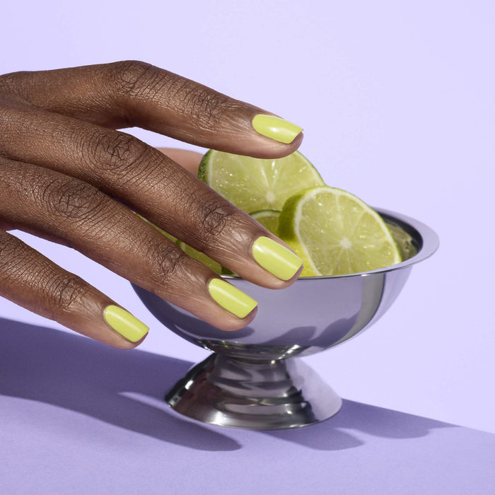 A hand with Manucurist Lime glossy green gel polish gently rests on a metal bowl filled with zesty citrus lime slices, set against a light purple background.