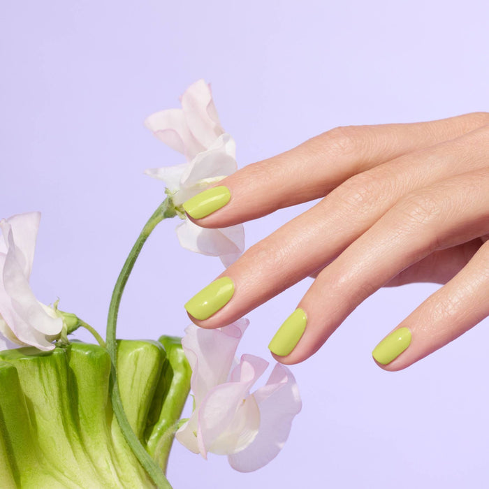 A hand with Manucurist Lime gel polish manicure touches delicate pale pink flowers against a pastel purple background.