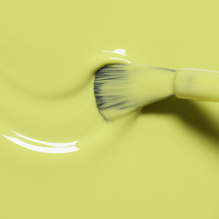 A close-up of a brush spreading Manucurist's Lime, a smooth, glossy anise green shade, over a yellow surface. Gentle curves and subtle texture highlight the nail polish’s zesty citrus vibrance.