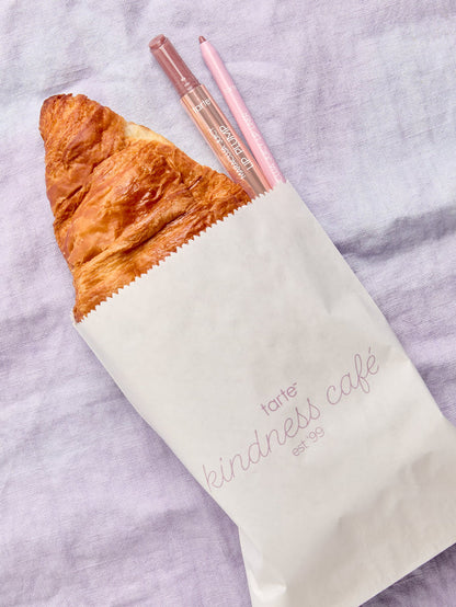 A croissant and two lip products from Tarte Cosmetics peek out of a paper bag labeled tarte kindness café est. '99, resting on a light purple fabric background.