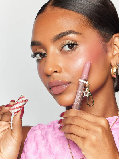 A person holds an open Tarte Cosmetics Maracuja Juicy Lip Plump product near their lips, displaying the applicator. They wear a pink textured top and gold hoop earrings, with a sparkling, candy-striped cap in the other hand.