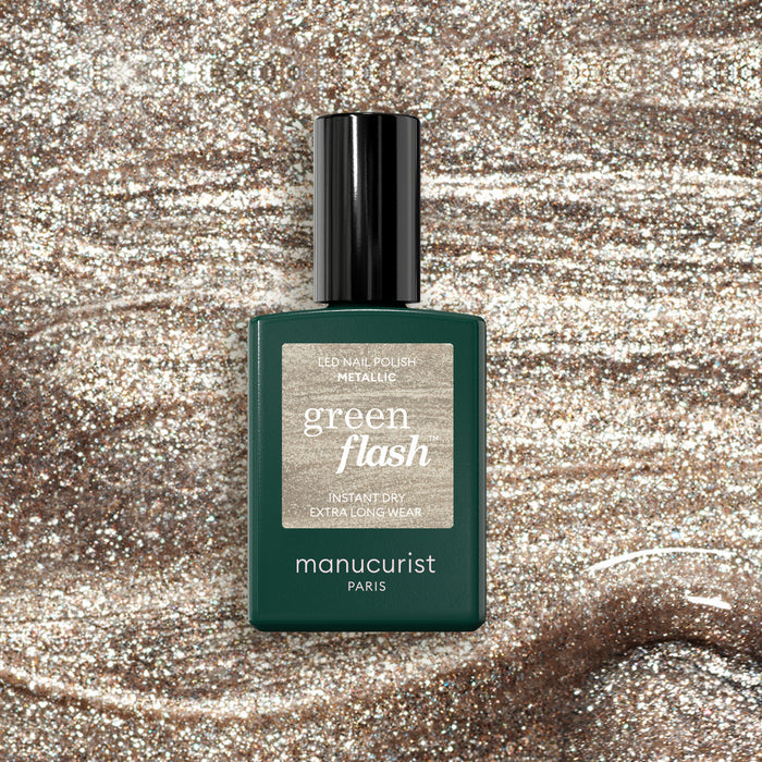 A bottle of Manucurist Metallic nail polish sits on a glittery surface that echoes the cool-toned gold shimmer and metallic highlights of the polish.