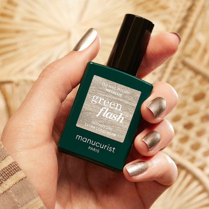 A hand with metallic silver nail polish and subtle highlights holds a bottle of Manucurist Metallic nail polish against a beige, textured background.