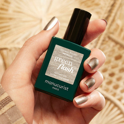 A hand with metallic silver nail polish and subtle highlights holds a bottle of Manucurist Metallic nail polish against a beige, textured background.