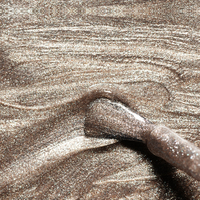 A close-up of a Manucurist Metallic nail polish brush applying shimmery, cool-toned gold and silver-brown polish with fine glitter and metallic highlights, creating a luxurious Green Flash effect on a smooth surface.