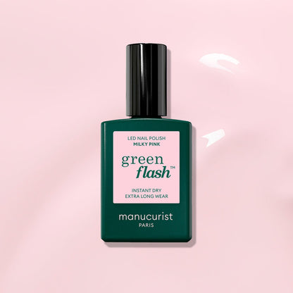 A bottle of Manucurist Milky Pink Green Flash gel polish, featuring a pink label on a dark green bottle with a black cap, rests on a light pink glossy surface.
