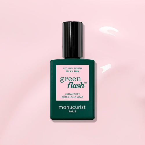 A bottle of Manucurist Milky Pink Green Flash gel polish, featuring a pink label on a dark green bottle with a black cap, rests on a light pink glossy surface.