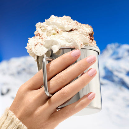 A hand with Milky Pink nails by Manucurist holds a metal mug of hot chocolate topped with whipped cream, set against snowy mountains and a clear blue sky.