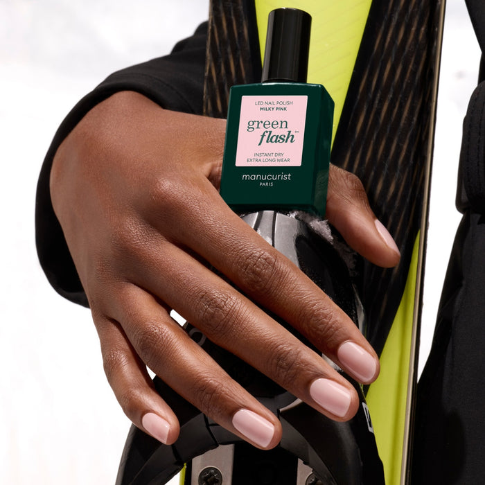 A hand with neatly manicured Milky Pink nails from Manucurist holds a green bottle of Green Flash gel polish against a black and yellow ski.
