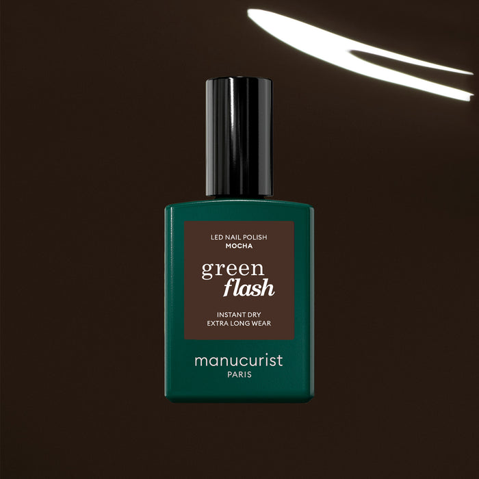 The Manucurist Mocha Green Flash LED nail polish, a dark brown shade inspired by exotic beans, is shown against a deep brown background with a white light streak. The bottle features a dark green body, black cap, and brown label.