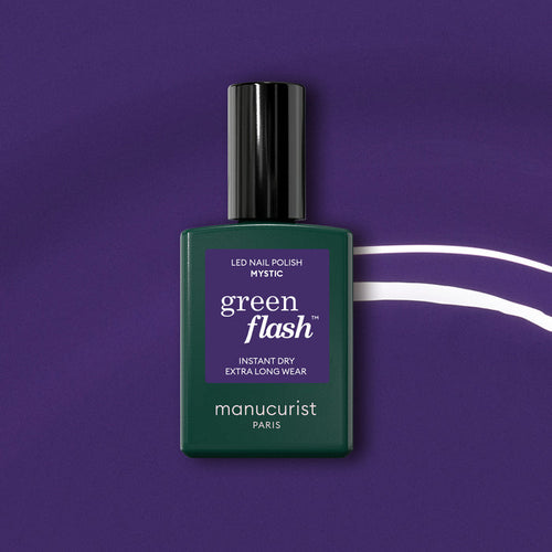A bottle of Manucurist Mystic Green Flash semi-permanent nail polish rests on a purple background with a wavy white line. The dark green bottle features a black cap and purple label, providing an opaque finish.