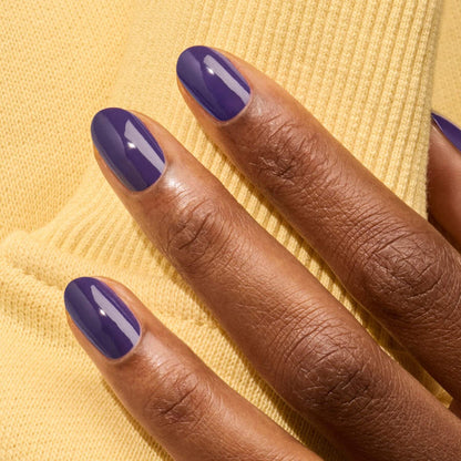 A hand with neatly manicured nails painted in intensely pigmented, glossy purple "Mystic" polish by Manucurist rests on soft yellow fabric.