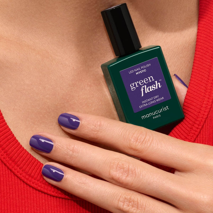 A hand with purple-painted nails, wearing a red top, holds a green bottle of Manucurist Mystic nail polish for an opaque finish.