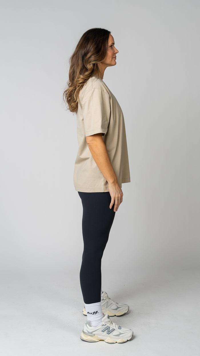 Woman wearing a beige t-shirt, black leggings, and white sneakers on a gray background. Side view