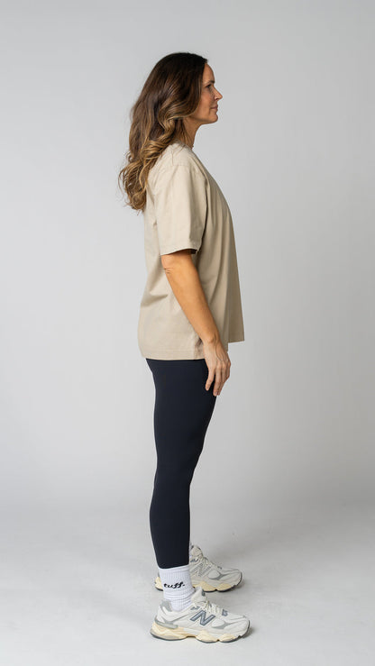 Woman wearing a beige t-shirt, black leggings, and white sneakers on a gray background. Side view