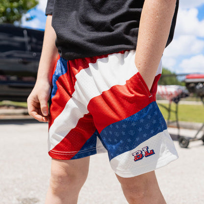 Lower body close-up of youth model in red white and blue Patriot Pitch shorts showing diagonal colorblock design and BL star logo at outdoor event