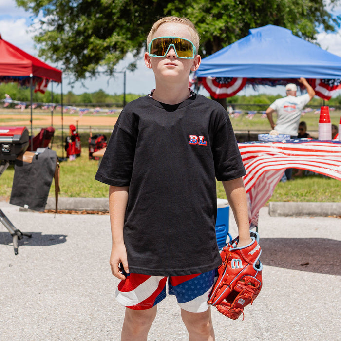 Front-facing youth male model wearing red white and blue Patriot Pitch shorts with black Patriot Pitch tee teal shield sunglasses and baseball glove at outdoor baseball event