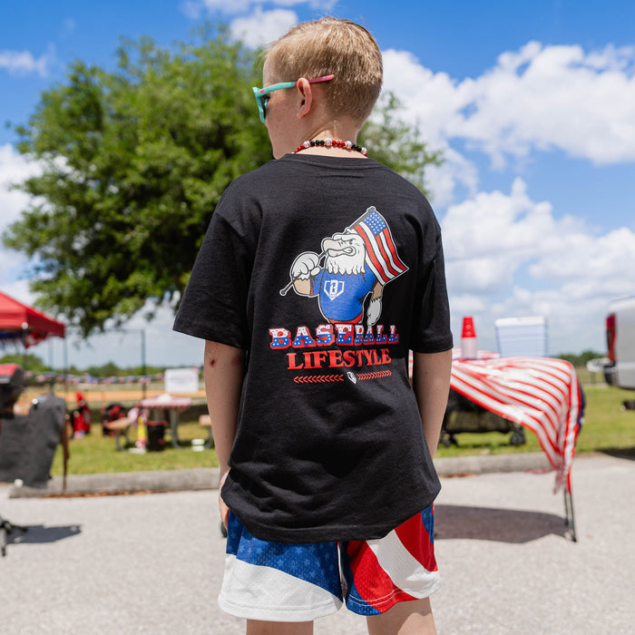 Back view of youth male model wearing red white and blue Patriot Pitch shorts and black Patriot Pitch tee showing Baseball Lifestyle eagle graphic at outdoor baseball event