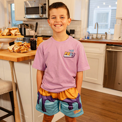 Young boy smiling in a kitchen wearing BL101 youth PB&J mesh shorts and a purple BL101 PB&J tee.