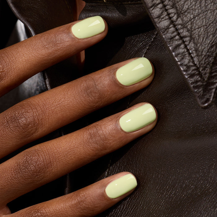 A close-up of a hand with short, neatly manicured nails painted in Pistachio by Manucurist, a pale green shade that softly glows, resting on a black leather jacket.