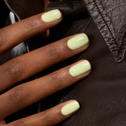 A close-up of a hand with short, neatly manicured nails painted in Pistachio by Manucurist, a pale green shade that softly glows, resting on a black leather jacket.