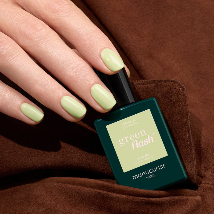A hand with neatly manicured nails painted in Manucurist Pistachio, a pale green LED Gel polish with yellow undertones, holds the bottle against a brown fabric background.