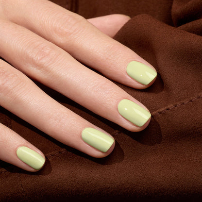 A close-up of a hand with neatly manicured nails painted in Manucurist's Pistachio, a pale green LED Gel polish with soft yellow undertones, resting on a brown fabric surface.