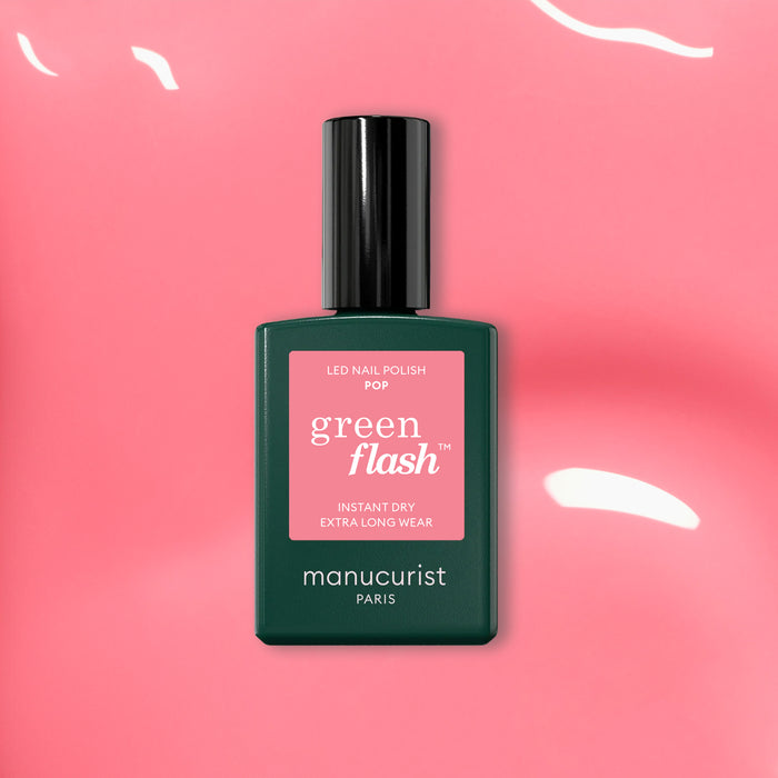 A bottle of Manucurist Pop Green Flash LED nail polish, a vibrant shade with orange undertones, rests on a glossy pink surface with reflected light, its black cap and green label standing out.